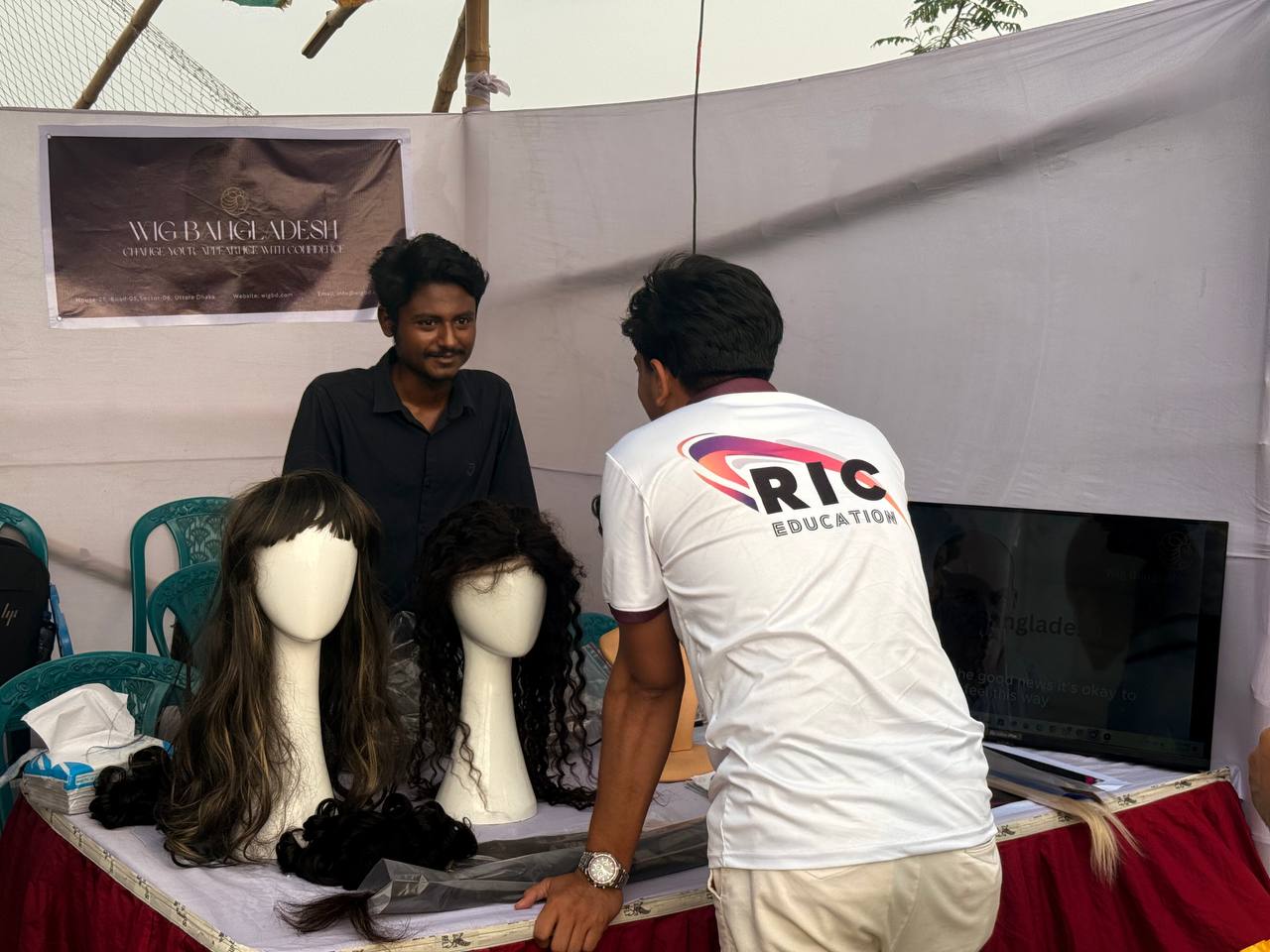 Students speaking with the Wig Bangladesh booth team beside displayed wigs