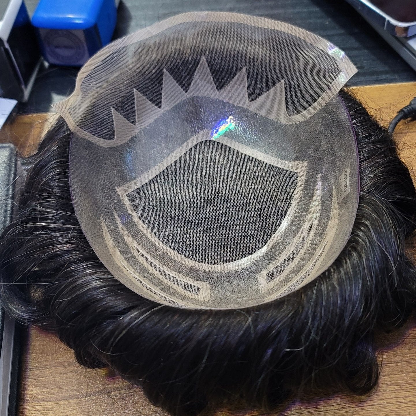 A black synthetic hairpiece with a clear mesh undercap and printed hairline guide, displayed on a wooden desk with a phone and other items in the background.