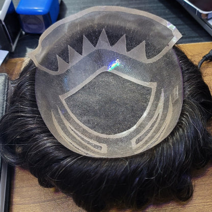 A black synthetic hairpiece with a clear mesh undercap and printed hairline guide, displayed on a wooden desk with a phone and other items in the background.